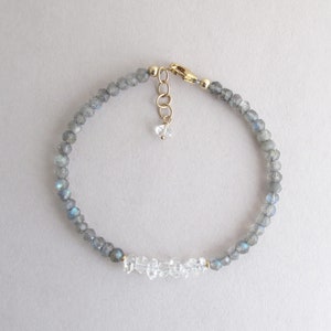 Pu&ograve; includere: Bracciale delicato con piccole perline grigie sfaccettate con riflessi blu, intervallate da accenti di cristallo trasparente. Il bracciale ha una chiusura e una catena di estensione color oro e un piccolo ciondolo di cristallo. Un gioiello semplice ed elegante.