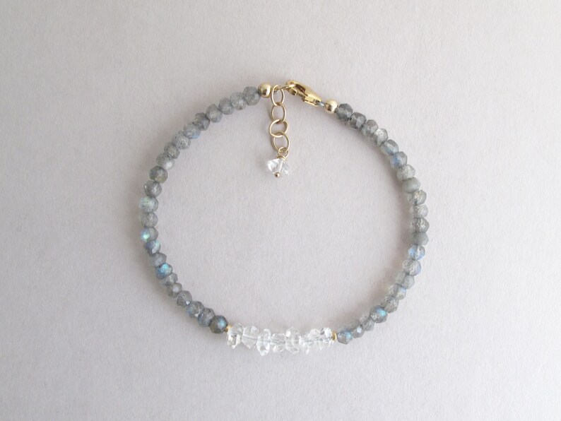 Pu&ograve; includere: Bracciale delicato con piccole perline grigie sfaccettate con riflessi blu, intervallate da accenti di cristallo trasparente. Il bracciale ha una chiusura e una catena di estensione color oro e un piccolo ciondolo di cristallo. Un gioiello semplice ed elegante.
