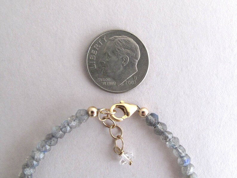 Pu&ograve; includere: Un delicato braccialetto di perline con piccole pietre preziose grigio-blu sfaccettate. Il braccialetto ha una chiusura color oro e un piccolo ciondolo di cristallo trasparente. Una moneta &egrave; inclusa per riferimento.