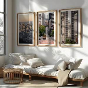 May include: Three framed landscape prints of San Francisco cityscapes. The prints feature a skyline view, a cable car on a steep street, and a building with a clock tower. The frames are light wood, and the images are displayed above a white couch.