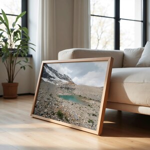 May include: A framed landscape photograph of a mountain range with a turquoise lake. The image is in a light wood frame and is leaning against a beige sofa. The scene includes a rocky terrain and a cloudy sky.