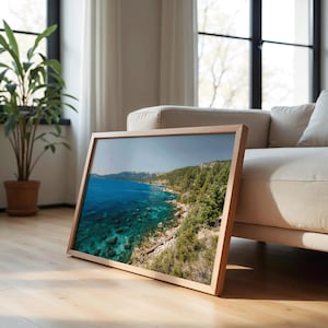 May include: A framed landscape photograph featuring a vibrant blue lake with clear turquoise shallows. The shoreline is lined with green trees and rocky terrain. The frame is made of light-colored wood, and the image is displayed indoors.