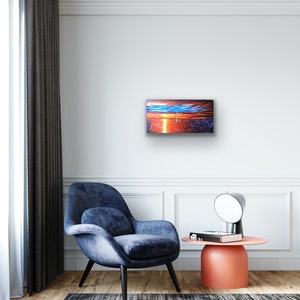 May include: A painting depicting a sunset over the ocean, with a sailboat visible in the distance. The sky is a blend of orange, red, and purple hues, while the water is a deep blue. The painting is framed and hangs on a white wall.