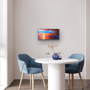 May include: A painting depicting a sunset over the ocean with a sailboat in the distance. The painting is framed and hanging on a white wall in a room with a white table and two blue chairs.
