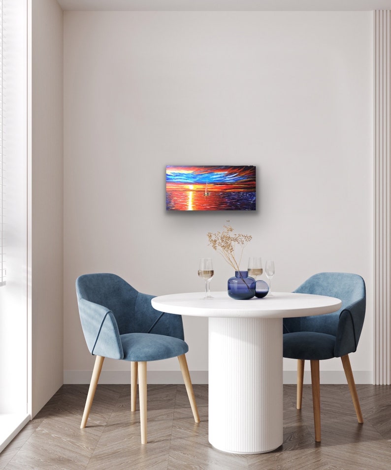 May include: A painting depicting a sunset over the ocean with a sailboat in the distance. The painting is framed and hanging on a white wall in a room with a white table and two blue chairs.
