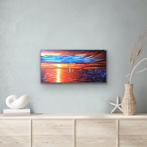 May include: A painting depicting a sailboat on a body of water at sunset. The sky is a blend of blue, orange, and red, with clouds. The water is a deep blue, reflecting the colours of the sky. The sailboat is white with a red sail.
