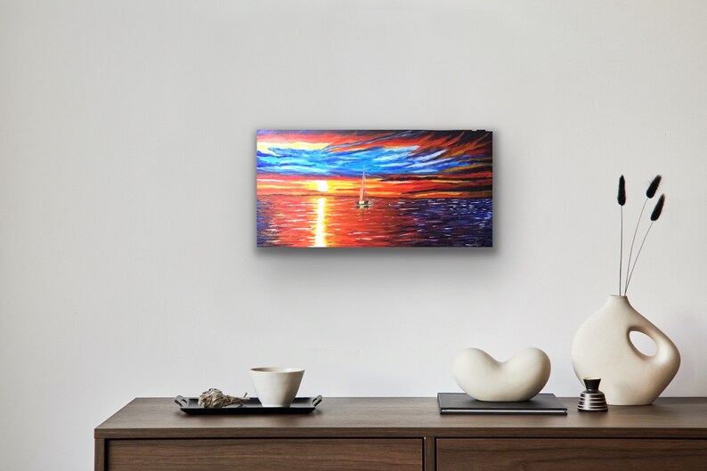 May include: A painting depicting a sailboat on the water at sunset. The sky is a blend of orange, red, and blue with clouds. The water is a dark blue with reflections of the sunset.