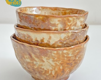 May include: A stack of three handmade ceramic bowls. The bowls have a rustic, textured appearance with a blend of brown and cream colors. The bowls are stacked on top of each other, showcasing their unique shapes and finishes. The text "CHA MORE" is in the upper left corner.