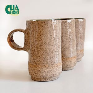 Może przedstawiać: Trzy nakrapiane ceramiczne kubki w odcieniach beżu i brązu. Jeden kubek ma uchwyt. Kubki mają złoty pasek u podstawy. Logo "CHA MORE" jest zielono-białe w lewym górnym rogu.