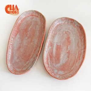 May include: Two oval-shaped ceramic serving platters. The platters have a reddish-pink glaze with a mottled, silvery-white interior. The rims are a darker red. The logo "CHA MORE" is in the upper left corner.