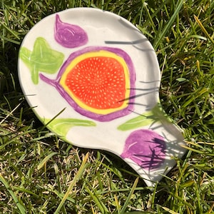 Op de afbeelding: Een witte keramische lepelhouder met een handgeschilderde illustratie van een doorgesneden vijg met een feloranje kern, paarse schil en groene bladeren. Het schaaltje heeft een onregelmatige, afgeronde vorm met een taps toelopend uiteinde.