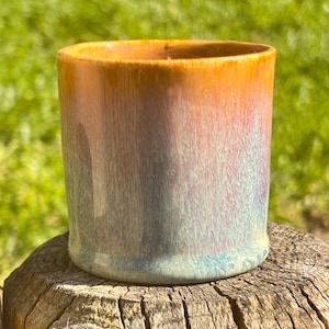 Puede incluir: Una taza de cerámica cilíndrica con un degradado de colores. La parte superior es naranja, pasando a morado y azul. La taza está sobre una superficie de madera desgastada, con un fondo verde borroso.