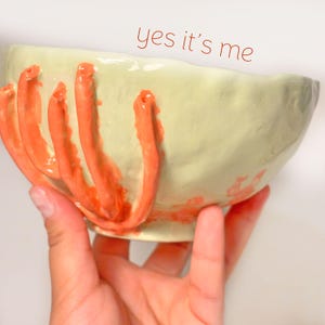 Puede incluir: Un cuenco de cer&aacute;mica hecho a mano con un esmalte verde claro y protuberancias anaranjadas en forma de tent&aacute;culos. El cuenco tiene el texto "yes it's me" en escritura roja. El cuenco est&aacute; sostenido en una mano.