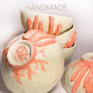 Puede incluir: Cuencos de cer&aacute;mica hechos a mano apilados, con base color crema y detalles texturizados en color coral. Los cuencos tienen una forma org&aacute;nica &uacute;nica. La palabra "HANDMADE" est&aacute; en la parte superior.