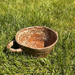 May include: A round, ceramic colander featuring a mottled brown and orange glaze with a glossy finish. The bowl has numerous small, evenly spaced drainage holes throughout its body and a single, sturdy, curved handle attached to the side.