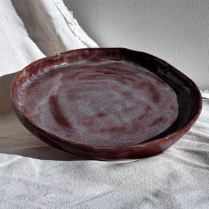 Könnte beinhalten: Eine handgefertigte, ovale Servierschale aus Keramik. Die Schale hat eine tief burgunderfarbene Glasur mit helleren, wirbelnden Tönen in der Mitte. Der Rand ist dunkler braun, und die Kanten haben eine organische, unebene Form.
