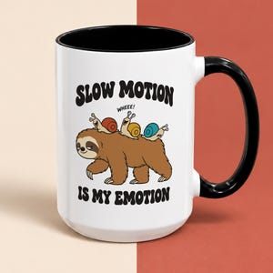 May include: A white ceramic mug with a black handle and interior. The front features a brown sloth with three colourful snails on its back. Above the sloth, the text reads SLOW MOTION and WHEEE!, and below the sloth, the text reads IS MY EMOTION.