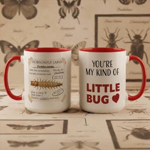 Op de afbeelding: Twee witte keramische mokken met rode handvatten en randen. Eén mok heeft tekst en een illustratie van een dobsonfly-larve. De andere mok zegt "You're my kind of little bug" met een hartsymbool. De mokken staan op een patroon.