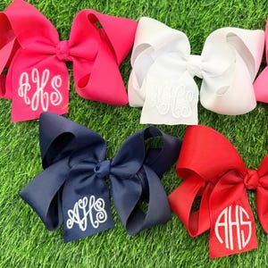 May include: Five large hair bows made of grosgrain ribbon in hot pink, white, light pink, navy blue, and red. Each bow features a centered, circular white monogram with the letters AHS embroidered on one of the bottom ribbon tails.