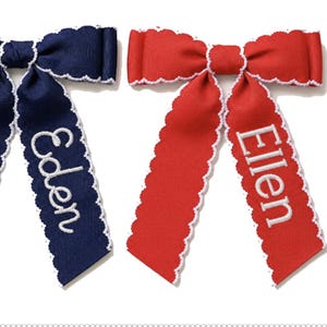 May include: Two hair bows with scalloped white trim edges. One bow is navy blue with the name Eden embroidered in white thread. The other bow is red with the name Ellen embroidered in white thread. Both bows feature long, dangling ribbon tails.