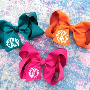 May include: Three large hair bows in teal, orange, and hot pink. Each bow is made of smooth ribbon with a knotted center and features a white circular monogram with stylized interlocking letters printed on one of the bottom ribbon loops.