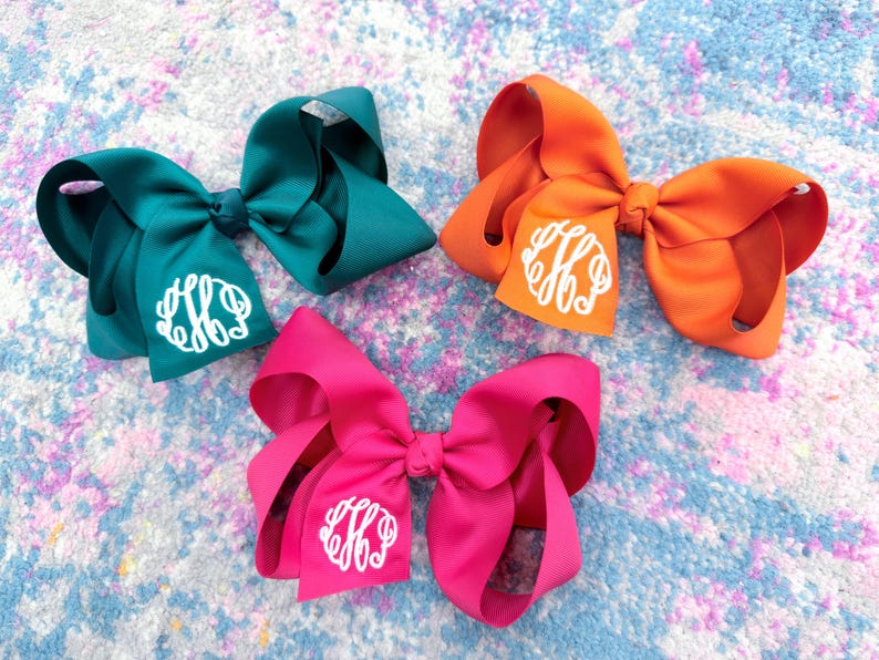 May include: Three large hair bows in teal, orange, and hot pink. Each bow is made of smooth ribbon with a knotted center and features a white circular monogram with stylized interlocking letters printed on one of the bottom ribbon loops.