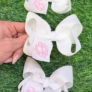 Three white hair bows made of ribbed grosgrain ribbon. Each bow features a centered, embroidered monogram in light pink thread.
