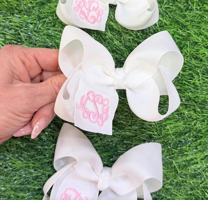 Three white hair bows made of ribbed grosgrain ribbon. Each bow features a centered, embroidered monogram in light pink thread.