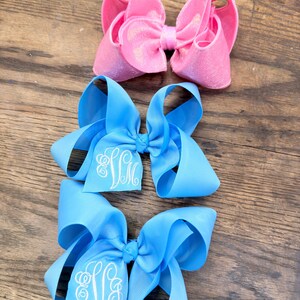 May include: Three hair bows arranged vertically. The top bow is a shimmering pink fabric. The middle and bottom bows are light blue grosgrain ribbon, each featuring a white embroidered monogram with the letters SM in a cursive script font.