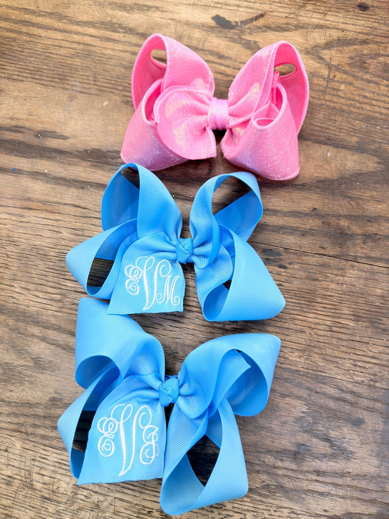 May include: Three hair bows arranged vertically. The top bow is a shimmering pink fabric. The middle and bottom bows are light blue grosgrain ribbon, each featuring a white embroidered monogram with the letters SM in a cursive script font.