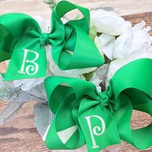 May include: Two bright green grosgrain ribbon hair bows featuring white embroidered letters. One bow displays the letter B and the other bow displays the letter P. The bows are constructed with multiple loops and a central knot detail on each piece.