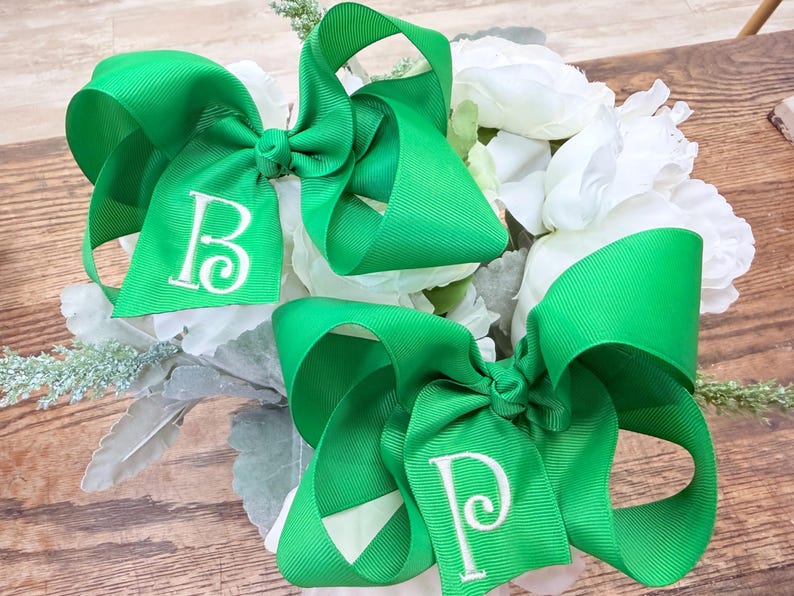 May include: Two bright green grosgrain ribbon hair bows featuring white embroidered letters. One bow displays the letter B and the other bow displays the letter P. The bows are constructed with multiple loops and a central knot detail on each piece.