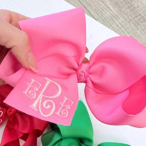 A large, bright pink hair bow made of grosgrain ribbon featuring a white embroidered monogram with the letters ERE in a cursive font. The bow has a structured, multi-looped design and is held up against a plain white background.