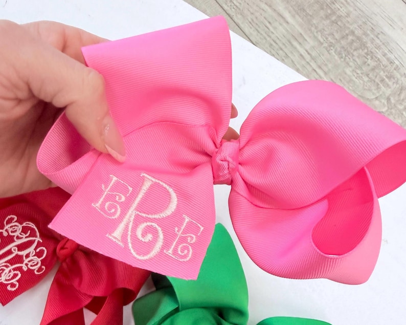 A large, bright pink hair bow made of grosgrain ribbon featuring a white embroidered monogram with the letters ERE in a cursive font. The bow has a structured, multi-looped design and is held up against a plain white background.