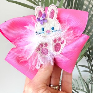 May include: A bright pink hair bow featuring a white bunny applique with pink ears and paws, blue eyes, and a purple bow. The bunny is surrounded by white feathers.