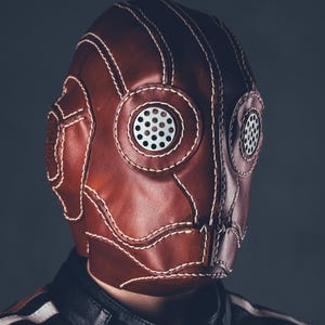 May include: A brown leather mask with white stitching and circular eye and mouth vents. The mask has a vintage, steampunk aesthetic. The mask is worn over the face and is part of a costume or cosplay.