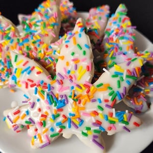 May include: A plate of leaf-shaped cookies covered in white icing and colorful sprinkles. The cookies are arranged on a white plate against a dark background. The sprinkles are a mix of red, yellow, green, blue, and purple.