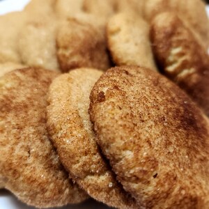 May include: A close-up of a plate of freshly baked cinnamon sugar cookies. The cookies are golden brown and have a slightly crispy texture. They are arranged in a pile, with some overlapping.