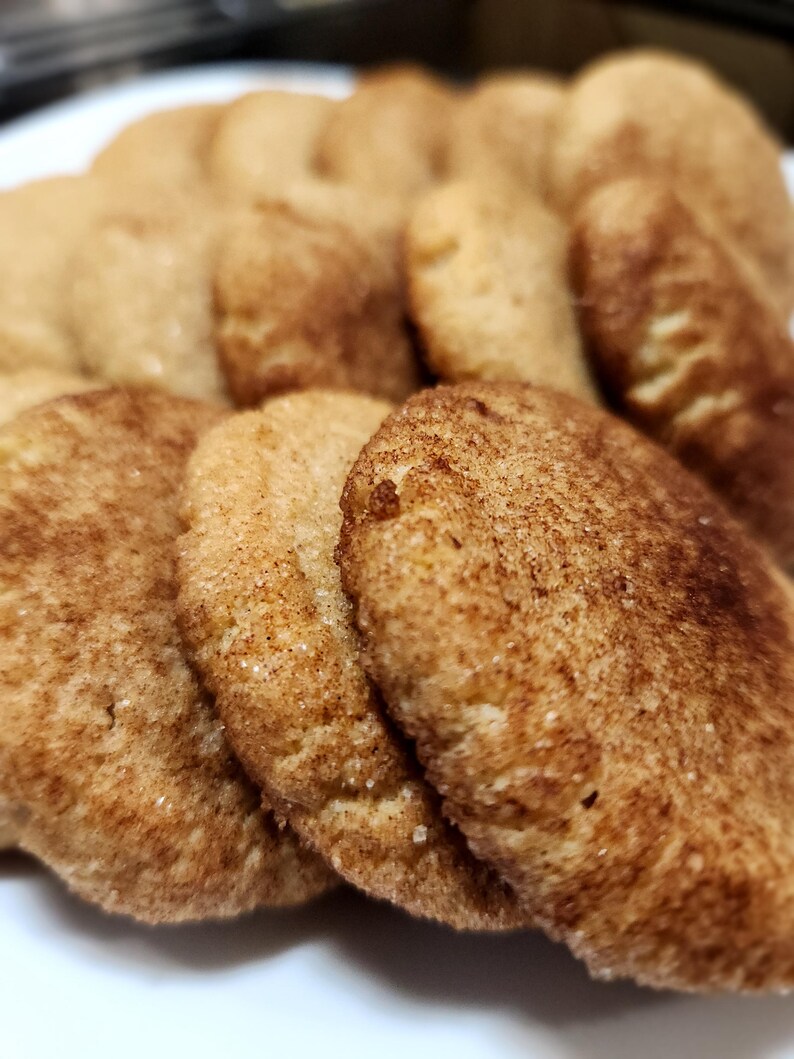 May include: A close-up of a plate of freshly baked cinnamon sugar cookies. The cookies are golden brown and have a slightly crispy texture. They are arranged in a pile, with some overlapping.
