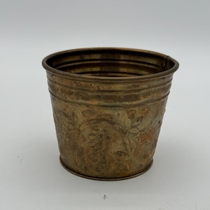 May include: A small, antique-style brass bucket with a floral pattern. The bucket has a cylindrical shape with a slightly flared rim and is likely used for decorative purposes. The brass has a warm, aged patina.