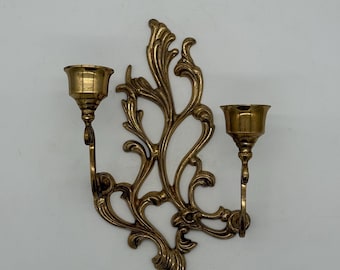 May include: A brass wall sconce with two candle holders. The sconce features an ornate, swirling design with a gold finish. The candle holders are positioned on either side of the decorative element.