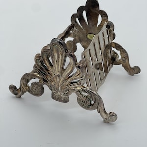 May include: Ornate silver-colored metal stand with a decorative shell-like design. The stand has a slotted back and curved legs, suitable for holding a small plate or dish. The stand is made in China.