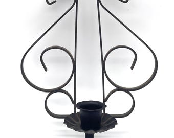 May include: A black wrought iron wall sconce. It has a candle holder with a scalloped base, decorative swirls, and a vertical design. The metalwork is detailed, adding a vintage touch to home decor. The sconce is designed for wall mounting.