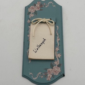 May include: A teal-colored wall hanging with a floral design featuring pink roses and green leaves. A small notepad with the word "Walberzy" is attached with a cream-colored string. The wall hanging has a decorative, curved shape.