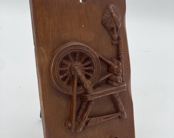 May include: A brown wooden wall hanging featuring a carved relief of a spinning wheel. The rectangular plaque has a hole at the top for hanging and is mounted on a black metal stand. The spinning wheel is intricately detailed.