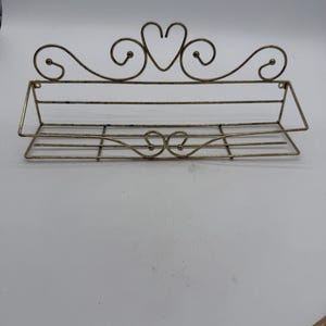 Può includere: Una mensola da parete decorativa in stile antico in metallo color oro. La mensola presenta un design a forma di cuore nella parte superiore e dettagli a volute. La mensola ha un cestello rettangolare per riporre oggetti.