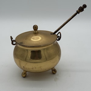 Puede incluir: Una pequeña olla redonda de latón con tapa y cuchara. La olla tiene tres patas pequeñas y dos asas. La tapa tiene un pequeño pomo en la parte superior. La cuchara es larga y delgada, con un mango decorativo. La olla y la cuchara son de latón brillante.