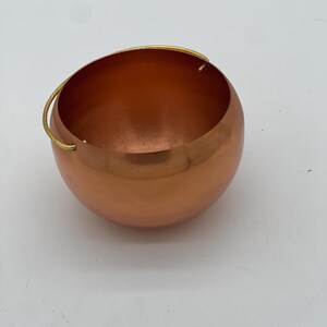 Puede incluir: Un cuenco esférico de color cobre con un asa dorada. El cuenco tiene una superficie lisa y reflectante y una forma redondeada. El asa está sujeta al borde superior del cuenco. El cuenco está vacío y se encuentra sobre una superficie blanca.