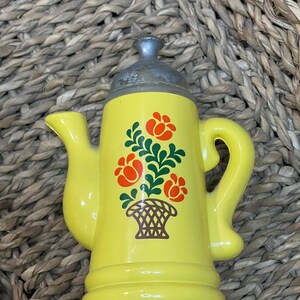 Puede incluir: Tetera de cerámica amarilla con tapa plateada y un diseño floral con flores rojas y hojas verdes en una cesta marrón.