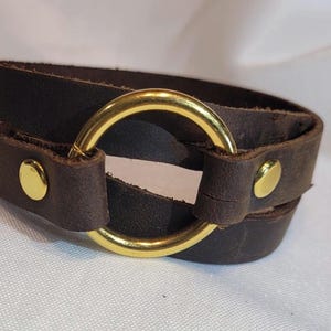 May include: Dark brown leather wrap bracelet with a gold-colored metal ring and gold-tone button studs. The bracelet wraps around the wrist twice. A stylish accessory for everyday wear.